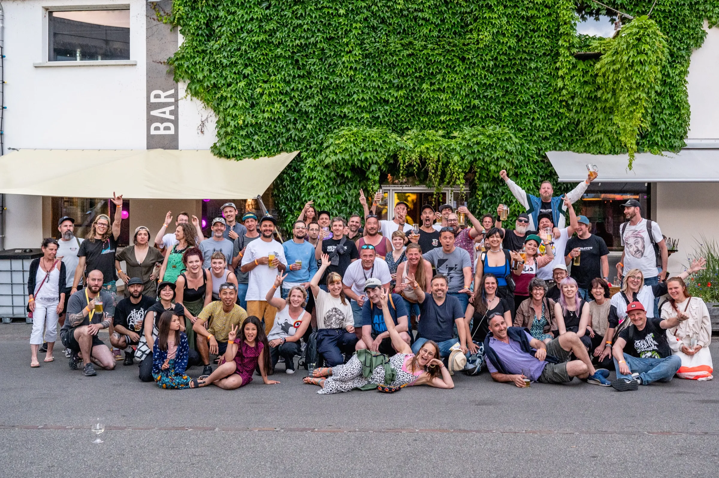 Rund sechzig Kunstschaffende und Mitwirkende des Festivals stehen und sitzen für ein Gruppenbild vor einer efeubewachsenen Hauswand